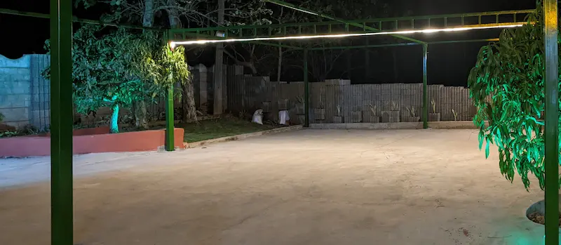 A concrete floored area with pillers holding up a led lighted line. Trees and grass are in the background.